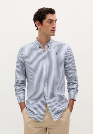 Blauw-wit gestreepte overhemddress met een button-down kraag, een logo op de borst en lange mouwen, gecombineerd met beige broek.