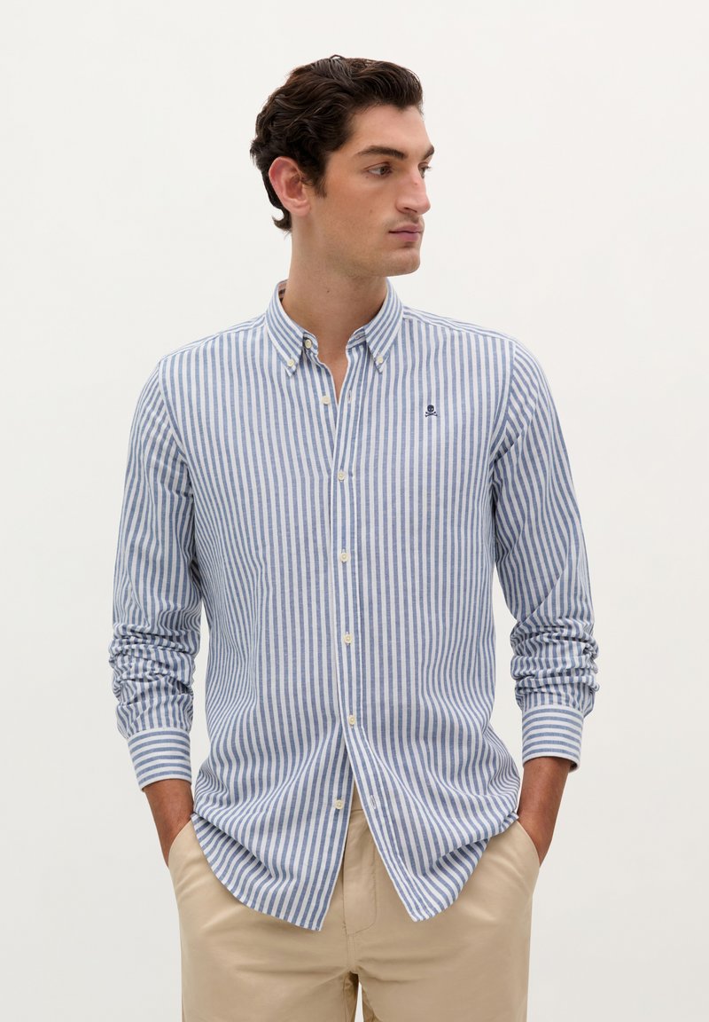 Blaue und weiße gestreifte Hemdbluse mit Button-Down-Kragen, Brustlogo und langen Ärmeln, kombiniert mit beigen Hosen.