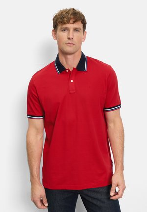 Mann trägt ein rotes Poloshirt mit marineblauem Kragen und Ärmelstreifen, steht vor einem schlichten weißen Hintergrund.