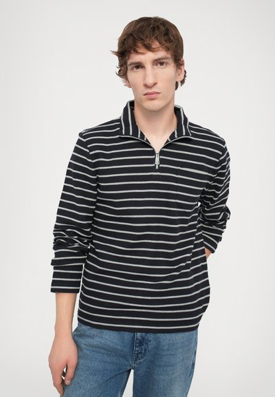 Polo cu dungi bleumarin și gri, cu mâneci lungi, guler cu fermoar la jumătate și un buzunar frontal, purtat cu blugi blue denim. Țesătură texturată.