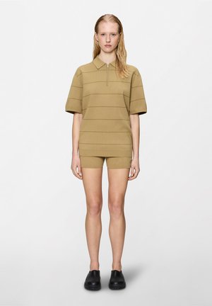 Femme aux cheveux blonds mouillés portant une chemise à manches courtes beige avec fermeture éclair, un short assorti et des chaussures noires sans lacets, debout devant un fond blanc uni.
