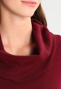 Maglione bordeaux con collo a scialle e trama a costine, caratterizzato da una vestibilità rilassata e un tessuto morbido che si adagia intorno al collo.