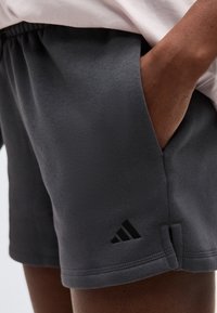 Κοντινό πλάνο μαύρων αθλητικών σορτς με εμπρόσθια τσέπη και ένα χέρι μέσα, με ένα μικρό μαύρο λογότυπο Adidas κοντά στο τελείωμα.