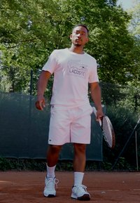 Vitt Lacoste T-shirt med logotyp, vita shorts och vita löparskor; håller en tennisklubba på en grusplan omgiven av grönska.