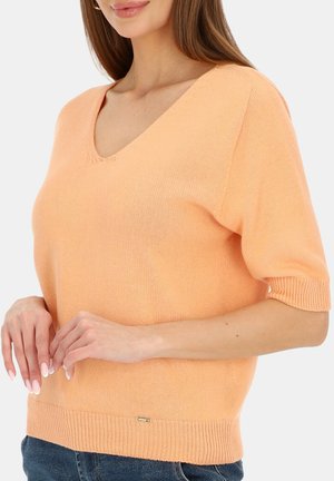 Kobieta ubrana w brzoskwiniowy, krótki sweter z dzianiny z dekoltem w serek i niebieskie dżinsy, z dłońmi delikatnie złożonymi na wysokości talii.