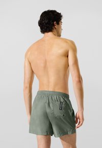 Póló nélküli férfi, göndör sötét hajjal, olívazöld fürdőnadrágban, hátoldalán zsebbel és logóval, háttal áll egy sima háttér előtt.