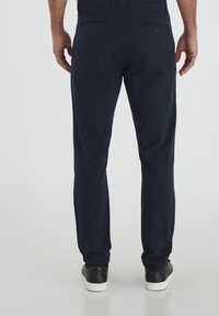 Chinos en coton bleu marine avec une coupe slim, dotés de deux poches arrière, d'une texture lisse et d'une fermeture par bouton. Portés avec des baskets noires.
