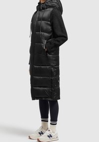 Długi czarny kamizelka puchowa z kapturem, teksturowane dzianinowe rękawy, boczne kieszenie i zapięcie na guziki u dołu. W połączeniu z białymi trampkami.