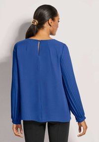 Blaue Bluse mit langen, gefältelten Ärmeln, rundem Ausschnitt und einer Schlüssel-Aussparung im Rücken. Texturiertes Material mit glatter Oberfläche.