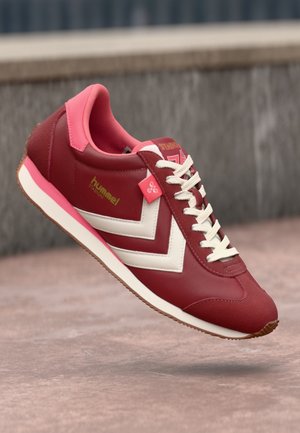 Roter und pinker Hummel Stadion Sneaker mit weißen Chevron-Streifen und Schnürsenkeln, gezeigt in der Luft über einer Betonfläche.