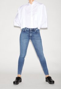 Valkoinen kauluspaita, jossa on puffeja hihnoissa, yhdistettynä sinisiin skinny-farkkuihin ja mustiin puureihin, joissa on hopeisia niittejä. Yksinkertainen ja puhdas muotoilu.