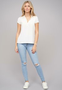 Top bianco a maniche corte con scollo a V e dettagli in pizzo sulle spalle, abbinato a jeans skinny strappati blu chiaro e sneakers bianche.