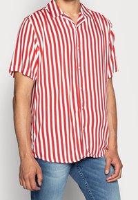 Camisa de manga corta con botones, con rayas verticales rojas y blancas, confeccionada en tela ligera, con cuello puntiagudo y corte holgado.