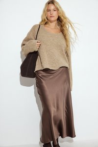 Beige Strickpullover mit lockerer Passform, kombiniert mit einem schokoladenbraunen Satinrock. Eine schlichte dunkle Handtasche rundet das Outfit ab.