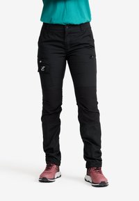 Person klädd i svarta cargo-byxor med knäskydd och rosa sneakers, står mot en vit bakgrund.