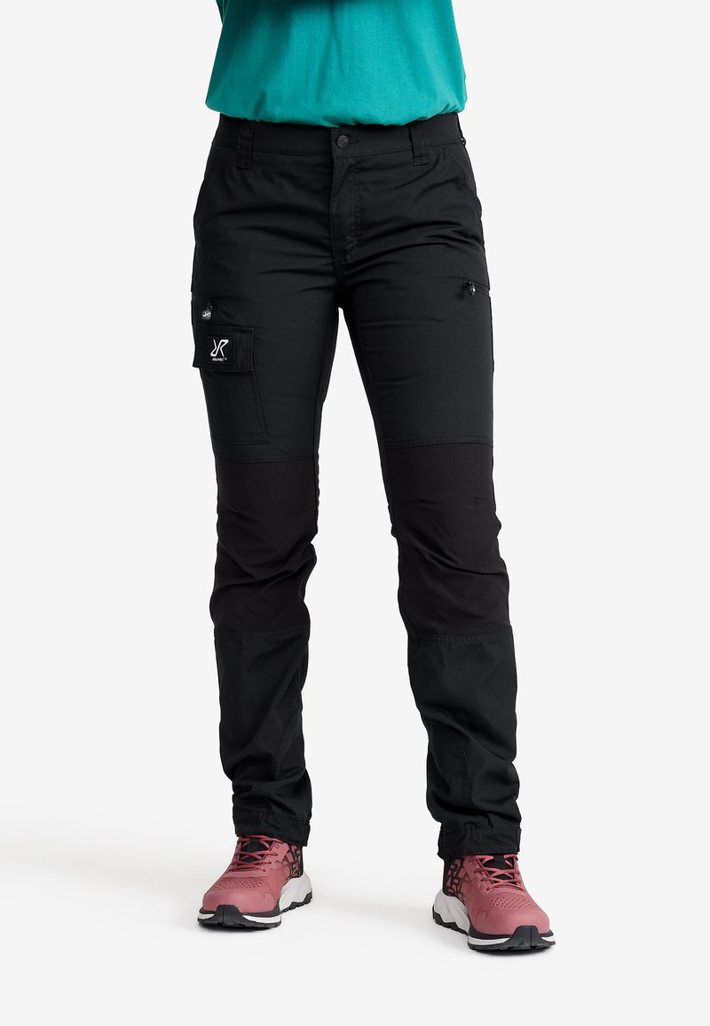 Person klädd i svarta cargo-byxor med knäskydd och rosa sneakers, står mot en vit bakgrund.
