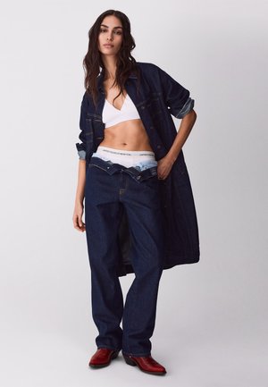 Kobieta w za dużym, ciemnym dżinsowym płaszczu i spodniach dżinsowych, rozpiętych, odsłaniających białą markową bieliznę, w białym biustonoszu i czerwonych butach, stojąca na jednolitym tle.