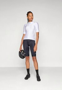 Biały dopasowany rowerowy krótki rękaw z zamkiem błyskawicznym, czarne rowerowe spodenki, czarne skarpety i buty. Trzyma czarny kask.