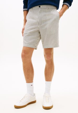 Man draagt een blauwe trui, beige en wit gestreepte korte broek, witte kousen tot aan de kuit en witte sportschoenen met bruine zolen, staand met handen in de zakken.