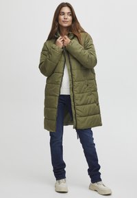 Olivově zelený nadýchaný dlouhý kabát s kapucí, se zapínáním na zip, patentkami a prošívanou strukturou. Kombinovaný s modrými džíny a béžovými teniskami.