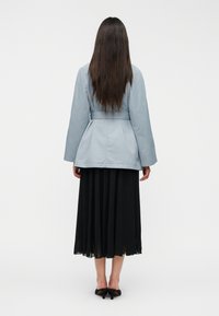 Φωτεινό γαλάζιο παλτό με ζώνη και μακριά μανίκια, σε συνδυασμό με μια ροή φούστα με πιέτες σε μαύρο χρώμα, εμφανισμένο από πίσω με φόντο λευκό.