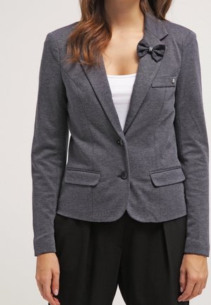 Kobieta ubrana w dopasowaną szarą marynarkę na dwa guziki, z kieszeniami z patkami, kokardą na klapie oraz czarne spodnie z wysokim stanem.
