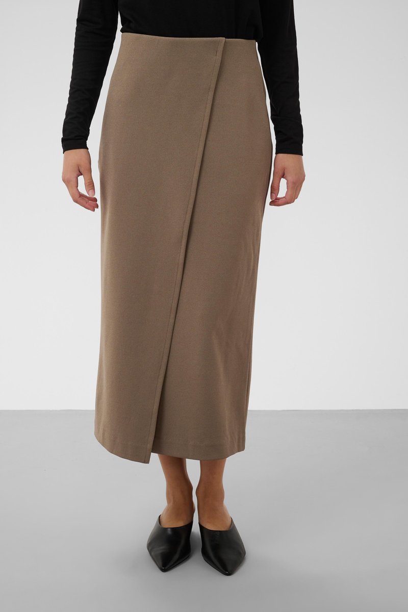 Kvinde iført en beige midi nederdel med omslag, sort langærmet top og sorte spidse sko står på en grå gulv.