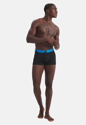 Atletische man die op blote voeten staat, gekleed in zwarte boxer briefs met een blauwe tailleband met Adidas-logo, op een effen witte achtergrond.