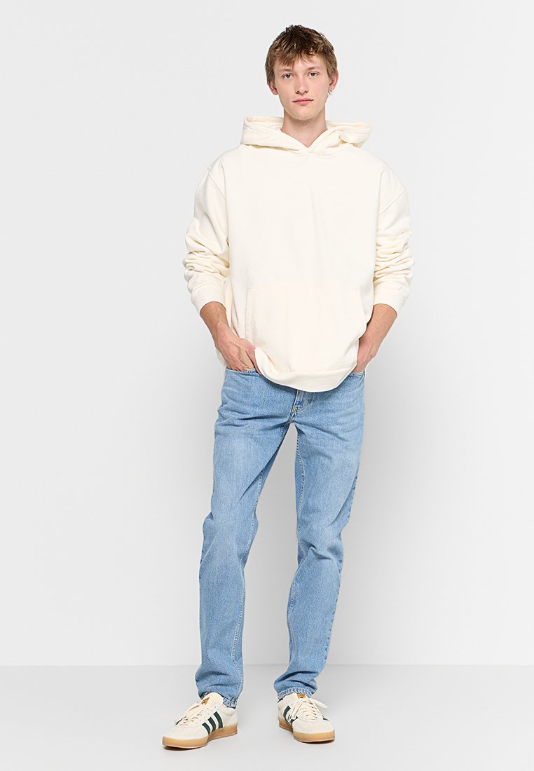 Crèmefarbene Hoodie mit Kängurutasche, weichem Material und verstellbarem Kordelzug, kombiniert mit hellblauen Jeans und beigen Sneakers.