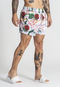 Shorts de bain blancs avec un motif floral et de chérubins vibrant, dotés d'une taille élastique et d'un cordon de serrage, assortis de sandales blanches.