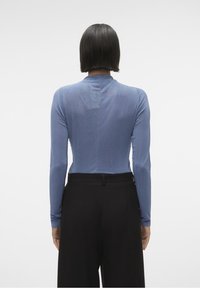 Pitkähihainen, vartalonmyötäinen paita pehmeässä sinisessä kankaassa, jossa on korkea kaulus ja hillitty tekstuuri, yhdistetty korkeavyötäröisiin mustiin housuihin.