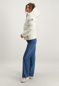 Witte pufferjack met capuchon, ritssluiting en zijzakken. Bevat een logo patch op de bovenarm. Gecombineerd met blauwe rechte jeans.