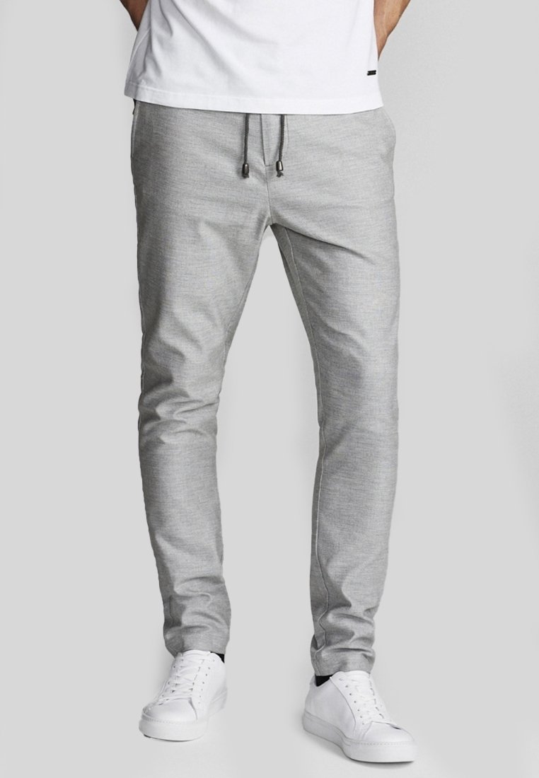 Pantalon de jogging gris avec taille à cordon, coupe fuselée, texture lisse, associé à des baskets blanches. Design simple, sans motifs visibles.
