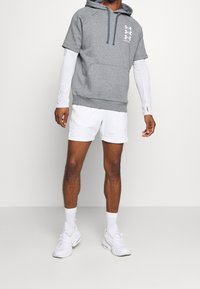 Hombre con sudadera corta gris sobre una camisa de manga larga blanca, pantalones cortos blancos, calcetas blancas y zapatillas blancas, contra un fondo blanco.