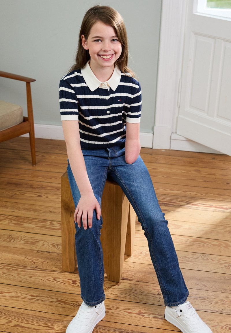 Polo shirt rayé bleu marine et blanc à manches courtes avec un col, jean en denim et baskets blanches. Assis sur un tabouret en bois.