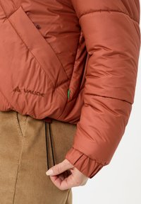 Roestkleurige pufferjack met gestikte ontwerp, elastische manchetten en verstelbare zoom. Bevat een logo en zijzak met ritssluiting.