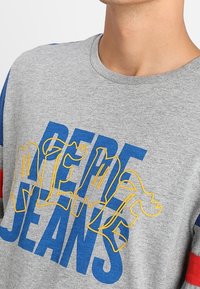 Šedé bavlněné tričko se silnými modrými a červenými pruhy. Na přední straně se nachází žlutý a modrý nápis "Pepe Jeans" v hravém písmu. Kulatý výstřih.