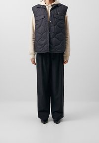 Chaleco negro acolchado con dos bolsillos delanteros, llevado sobre una sudadera crema. Pantalones negros holgados y zapatos de punta completan el conjunto.