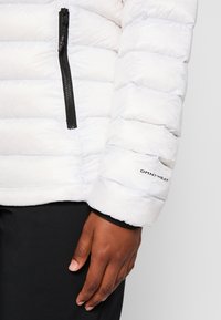 Vit puffjacka med en vertikal svart dragkedja, horisontell quiltad design och en Omni-Heat-lapp på ärmen. Svart inre foder.