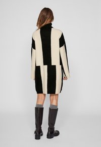 Robe-pull à motif noir et crème avec un col montant, une coupe oversize et une texture côtelée, s'étendant jusqu'au milieu des cuisses, associée à des bottes montantes jusqu'aux genoux.