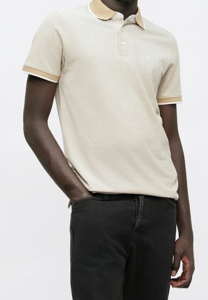 Homme portant un polo à manches courtes beige avec bordure beige clair et pantalon noir, debout avec une main dans la poche sur un fond blanc.