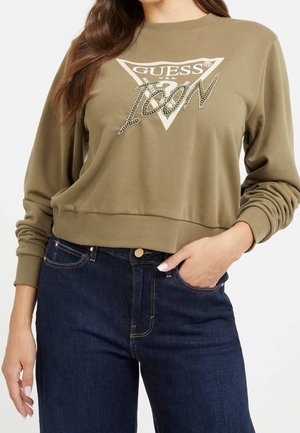 Femme portant un sweat-shirt olive vert de la marque Guess avec "ICON" en strass et un jean bleu foncé, une main dans la poche, sur un fond blanc.