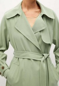 Světle zelený trench coat s páskem, širokými klopami, bo�čními kapsami a knoflíkovými manžetami, nošený přes béžový top.