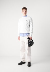 Sudadera blanca con logo, camisa de rayas azules debajo, pantalones crema, zapatos de cuero negros, sujetando un pequeño bolso negro.