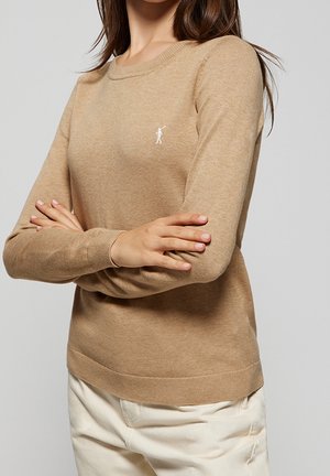 Vrouw draagt een zandkleurige trui met lange mouwen en een klein wit polo-speler logo, armen gekruist, gecombineerd met off-white broek.
