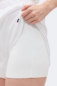 Χέρι σηκώνει λευκά εξωτερικά σορτς για να αποκαλύψει εφαρμοστά λευκά συμπιεστικά σορτς με ορατά ραφές και μερικό μαύρο λογότυπο.