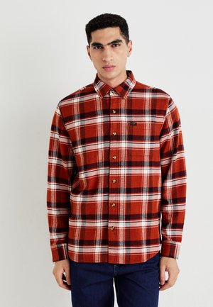 Chemise en flanelle à carreaux rouges et noirs avec un col boutonné, présentant une petite étiquette avec logo et des boutons marron, tissu texturé.