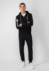 Sudadera negra con el logo blanco de Champion en la manga, combinada con pantalones de chándal negros. El modelo lleva zapatillas blancas, de pie con los brazos cruzados frente a un fondo simple.