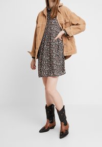 Chaqueta de pana marrón sobre un vestido negro de flores. Botas de vaquero marrones y negras con estrellas y detalles recortados. Fondo neutro.