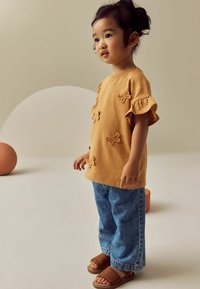 Senf-farbenes Kurzarmoberteil mit floraler Stickerei, kombiniert mit blauen Jeans und braunen Sandalen. Einfacher, lässiger Schnitt mit Rüschensaum.
